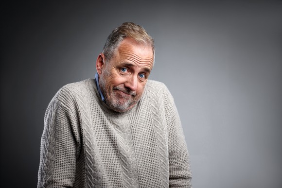 An older man in a gray sweater, shrugging his shoulders.