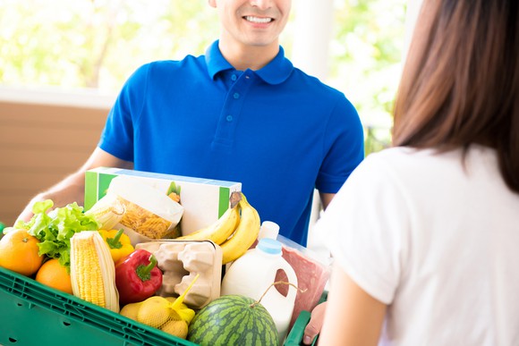 A smiling man in a blue shirt carrying a box full of fresh produce and milk and about to hand it to a woman customer