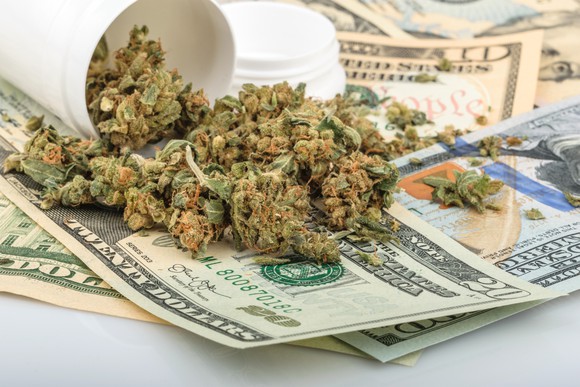 A tipped over bottle containing dried cannabis buds lying on a messy pile of cash.