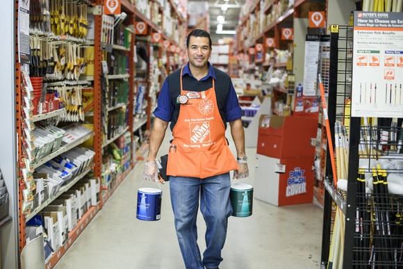 Home Depot associate carries two buckets of paint down store aisle.