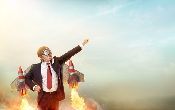 A businessman wearing a suit and strapped to metal wings with rockets prepares to lift off into the sky.