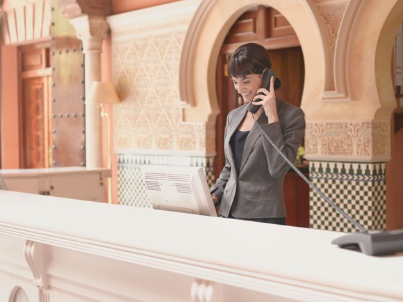 Hotel receptionist smiling and talking on phone at ornate reception desk.