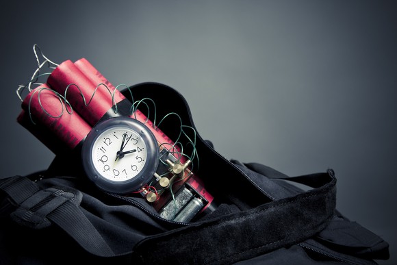 Black backpack holding sticks of red dynamite with a small clock attached to it.
