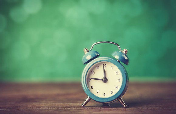 An alarm clock against a green background, sitting on a wooden surface.