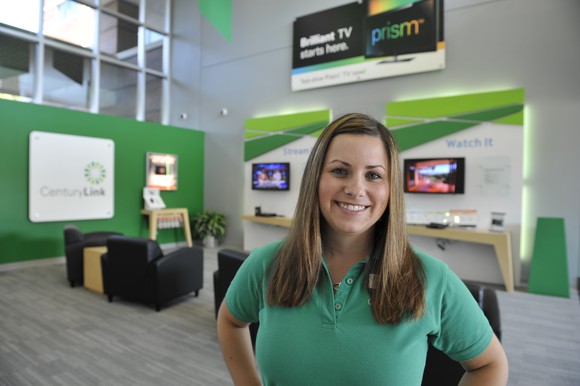 A woman in a green shirt in front of a CenturyLink and Prism TV display.