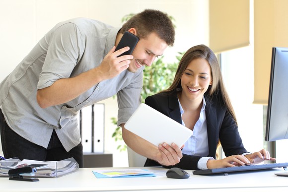 At the office, two young businesspeople communicate using a tablet, a phone, and a computer.