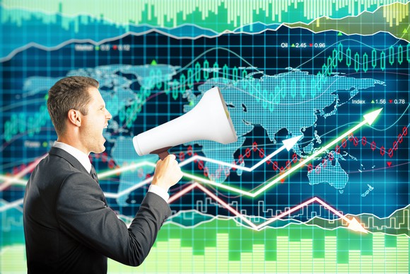 A man yells into a megaphone in front of a background showing ascending stock price charts and a map of the world.
