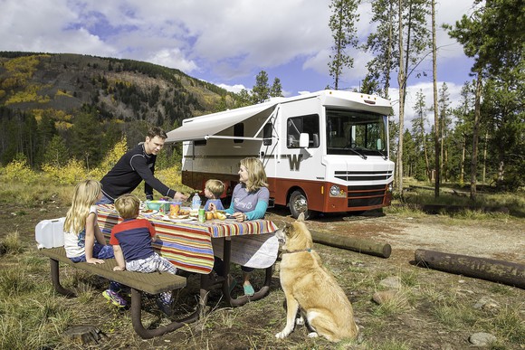 Two adults and two kids at a picnic table at a campsite, with a dog nearby, and a Winnebago RV parked in the background.