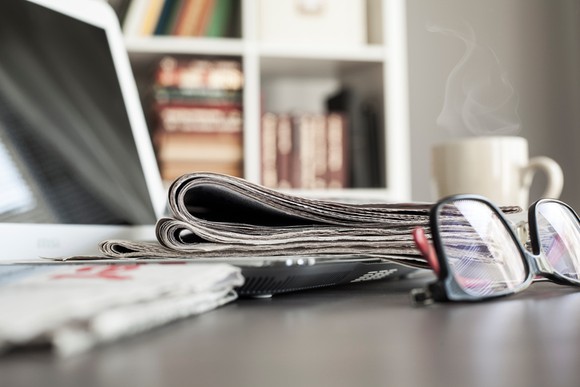 Close-up of a laptop, steaming cup of coffee, pair of glasses, and newspaper on desk.