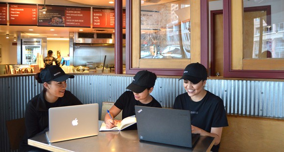 Chipotle employees sitting around a booth at a Chipotle using computers and writing in a notebook.