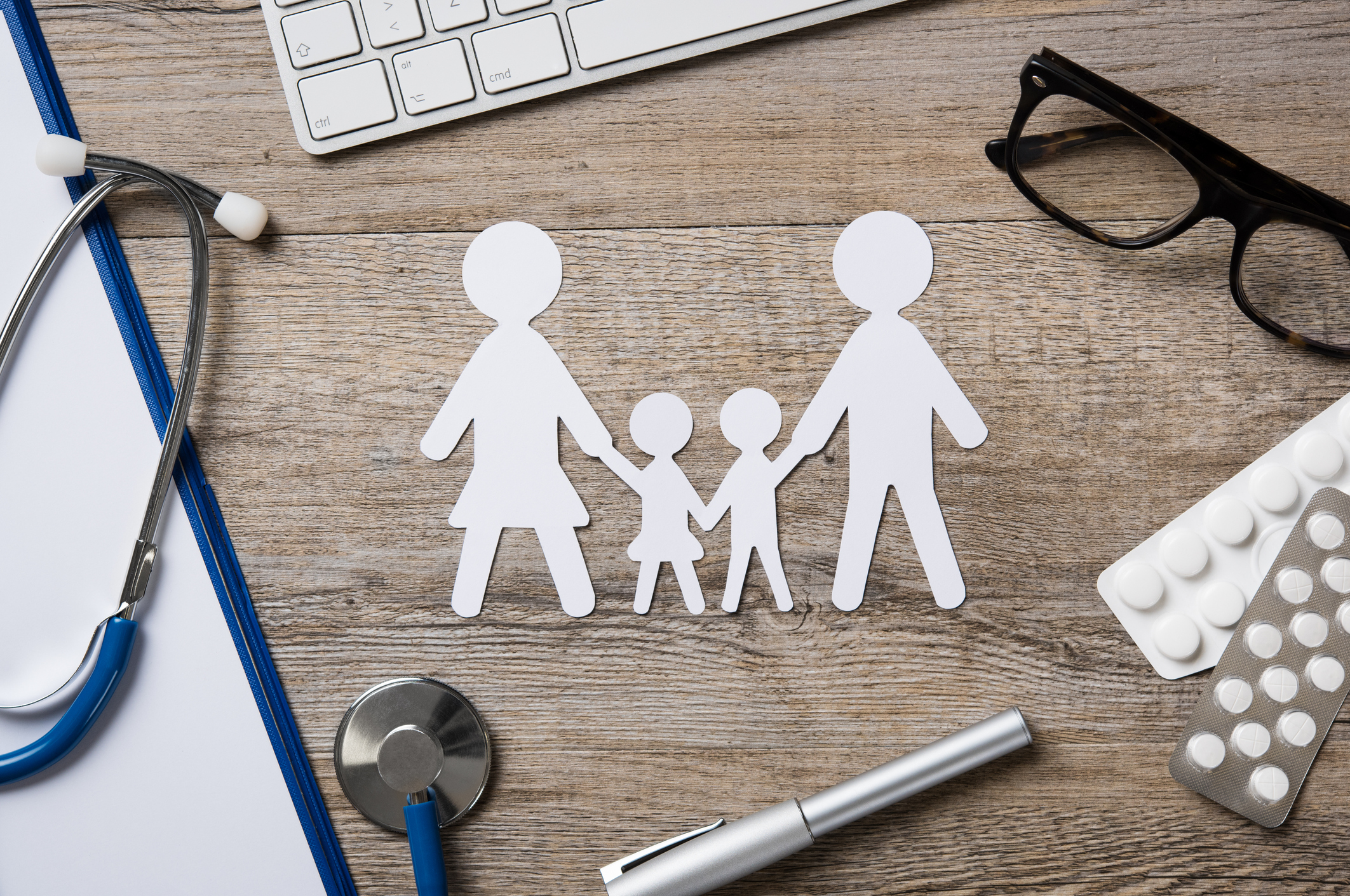 A paper cut out family rests on a desk amidst pills, glasses, a stethoscope, and a pen.