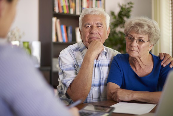 A senior couple with serious expressions look across a desk at a man in an office setting.