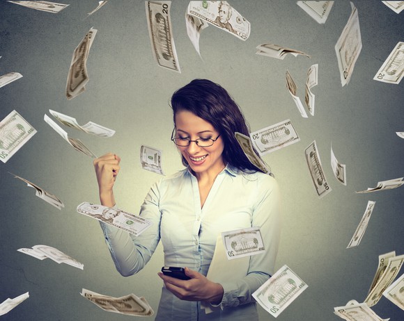 A woman pumping her fist as she checks her phone and cash money falls around her.