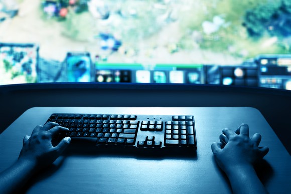 Hands on computer keyboard and mouse with PC monitor displaying a game in the background.
