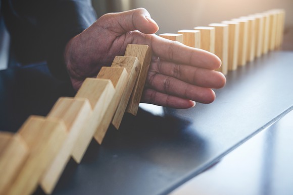 A man's hand prevents a series of falling wooden block from knocking over a series of upright wooden block