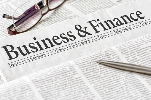 Close-up of the business and finance section of a newspaper, with a pen and a pair of glasses.