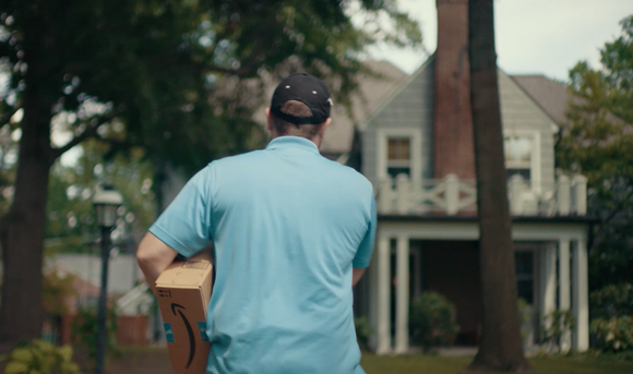 Back view of an Amazon delivery man carrying an Amazon package while waling toward a house in a suburban area.