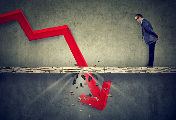 A man in a suit looks down at a falling red arrow breaking through a concrete floor.