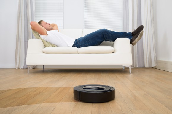 A man reclines on a white couch while a robotic vacuum cleans the floor.