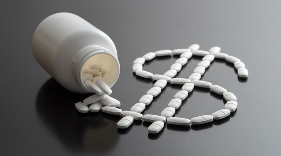 A pill bottle on its side with pills spilling out in the shape of a dollar sign.