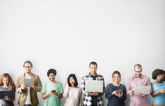 A group of people standing against a white wall using smartphones, tablets, and laptops.