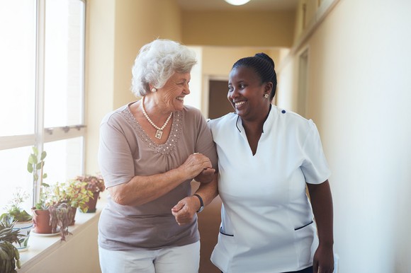 An older woman smiles and walks arm-in-arm with a healthcare provider.