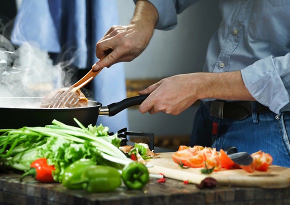 A chef prepares hotpot items on a stove top frying pan at home, with various vegetables on a chopping board on top of the stove.