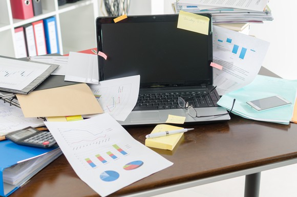 A cluttered desk with papers, a calculator, a stack of Post-its with a pen on top, and a smartphone around it, as well as a pair of glasses on top of it.