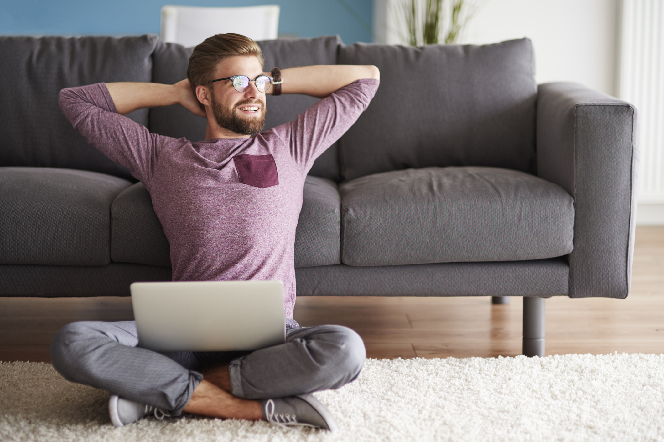 A smiling, bespectacled man sitting on the floor with leg crossed, arms up behind his head, and a laptop on his lap