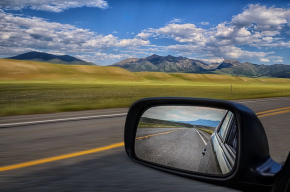 The countryside as viewed from the side window of a car. The side mirror of the car is shown in the foreground.