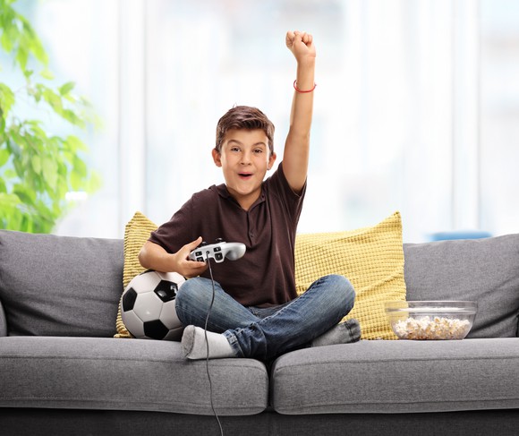 A smiling boy sitting on a couch holding a video game controller with his fist raised in the air.