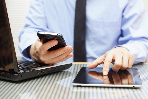 A businessman at his desk, working a latop, a smartphone, and a tablet all at once.