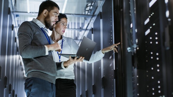A man holds a laptop in his left hand while a woman stands next to him pointing with her left hand at servers.
