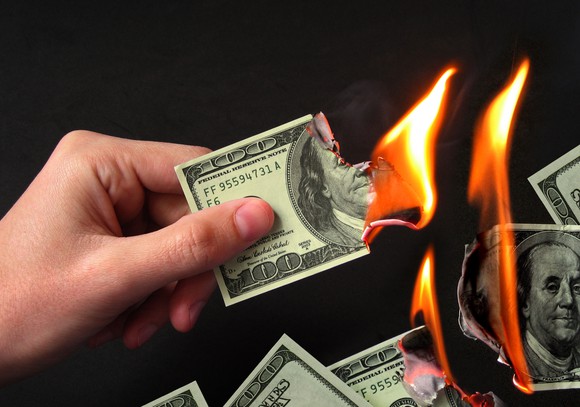 Hand holding a burning $100 bill with other bills burning in the background.