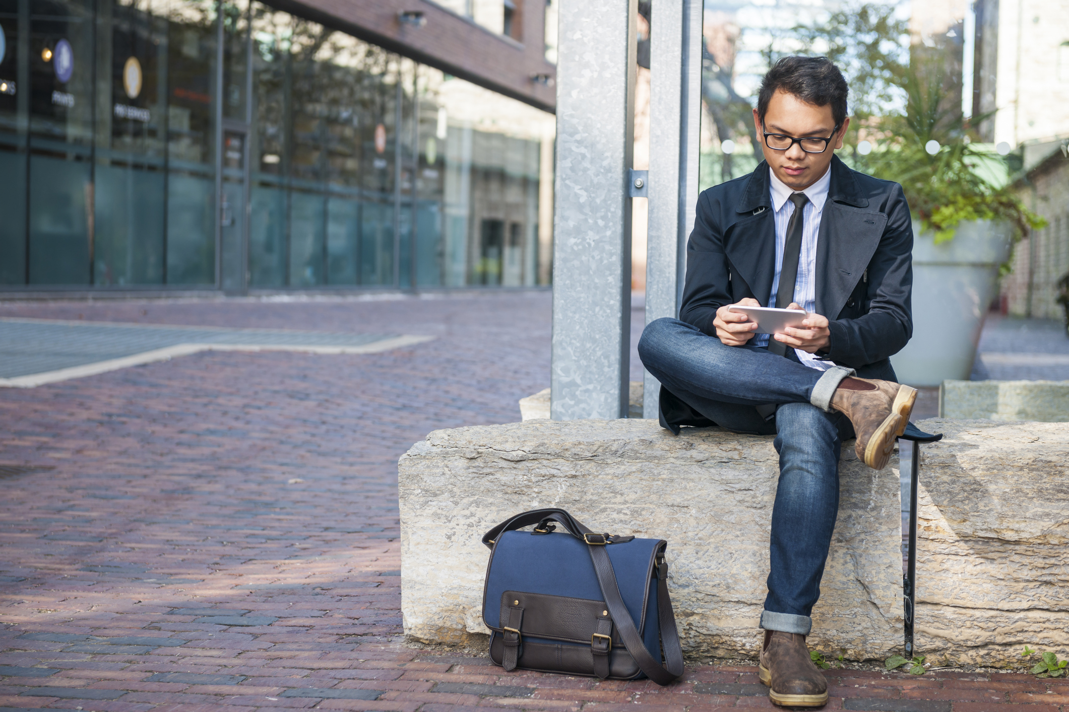 A bespectacled man in jeans and a suit jacket with his blue messenger bag on the ground as he checks his mobile phone while seated on a concrete bench outdoors