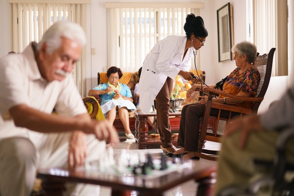 A doctor tends to an older woman inside an assisted living facility, as a game of chess is being played in the foreground
