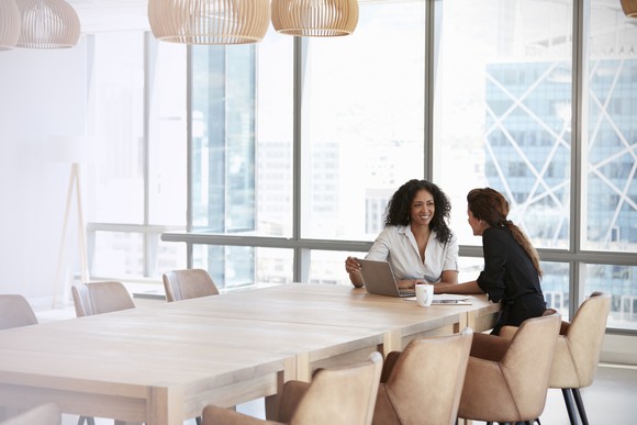 Two businesswoman talking at the end of a conference room table.