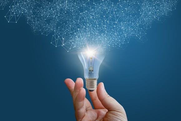 Hand holding a lit light bulb whose top has dissolved into interconnected points of light.