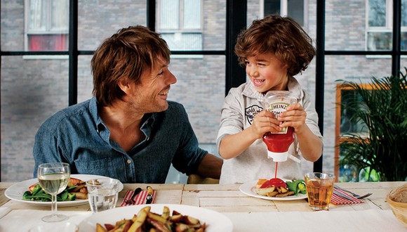 Child squeezing Heinz Ketchup bottle over food while smiling at father at dinner table.