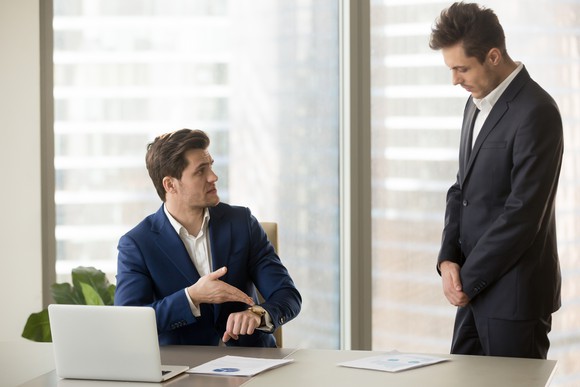 Professional man at desk points at his watch while another professional man stands nearby with his head down