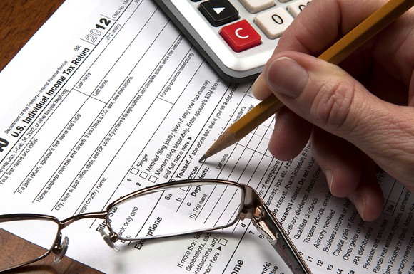 Hand holding a pencil, filling out a paper tax return beside glasses and calculator.