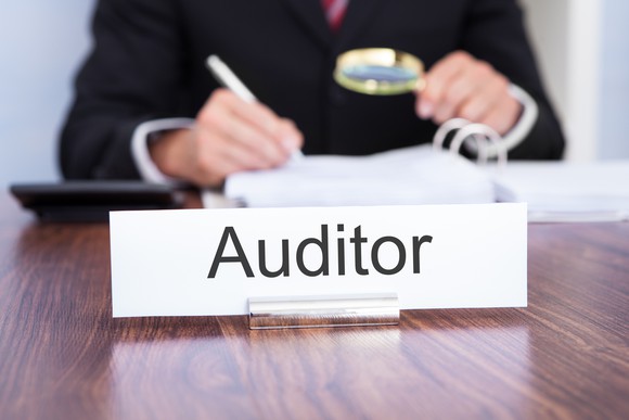 Man with magnifying glass looking over a document with an auditor sign in front of him