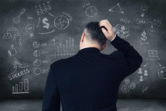 A man in a suit scratches his head while staring at a chalkboard covered by diagrams.