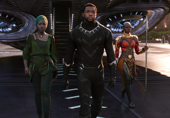 Chadwick Boseman in a screenshot as the superhero Black Panther, walking out of a spacecraft with women on each side of him.