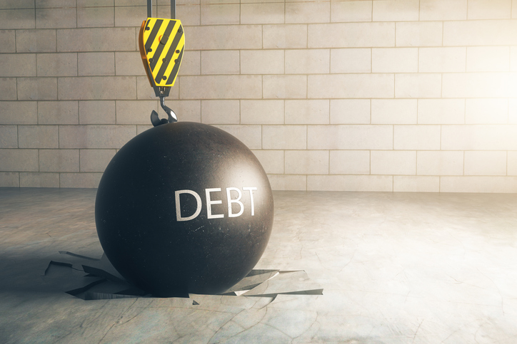 A black wrecking ball, with the word DEBT written on it, on top of a floor that it has obviously broken upon landing