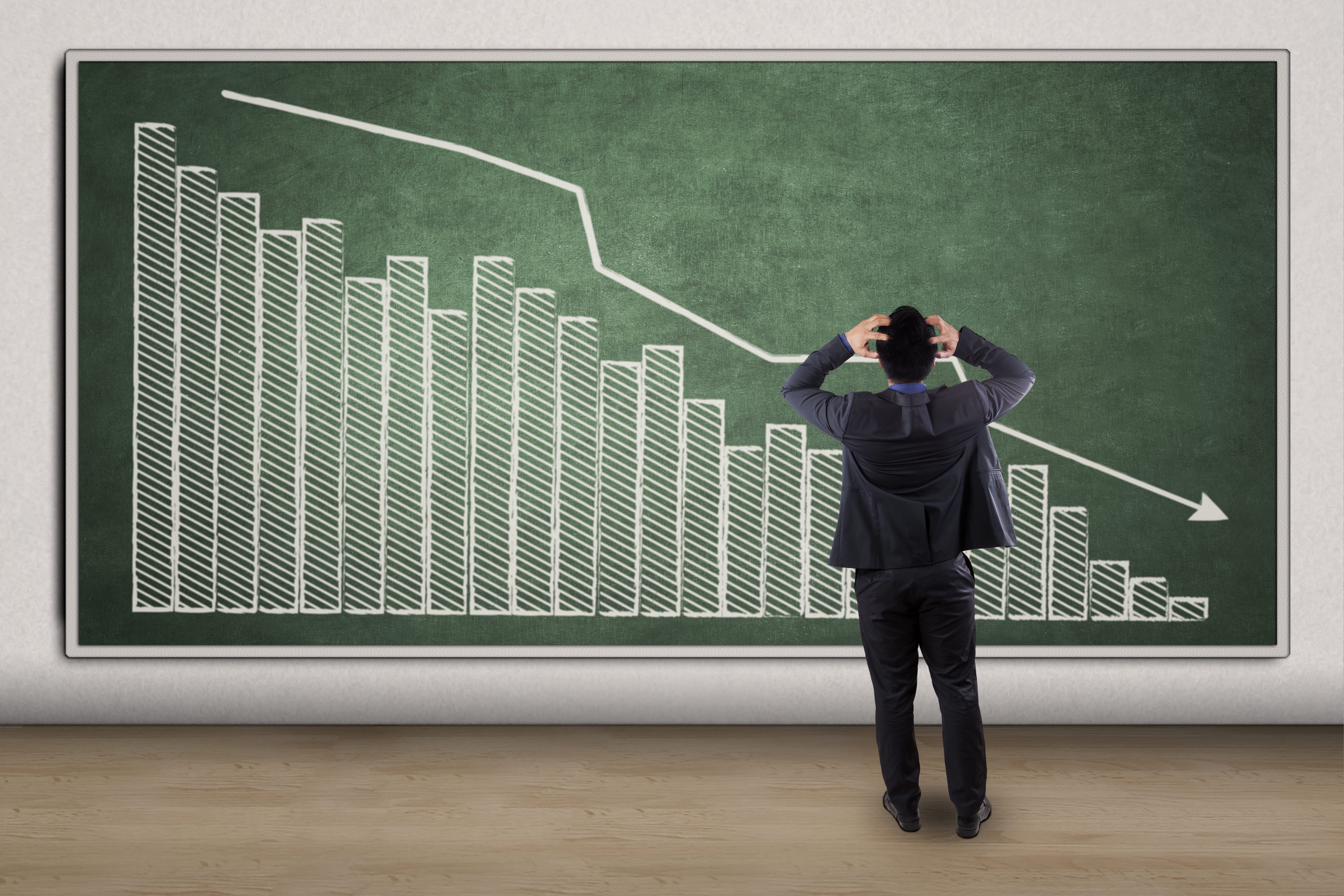 A graph in descending order, with an arrow pointing down, on a green chalkboard as a man in front of it clutching his head