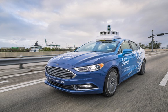A blue and white Ford Fusion with visible self-driving sensor hardware on a Miami street.