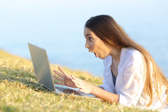 Young woman with mouth agape while looking at a laptop screen that's set on green grass.