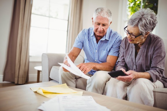 A senior couple sits on a sofa, with papers spread out on a table in front of them. The man holds a document and points to it while the woman uses a calculator.