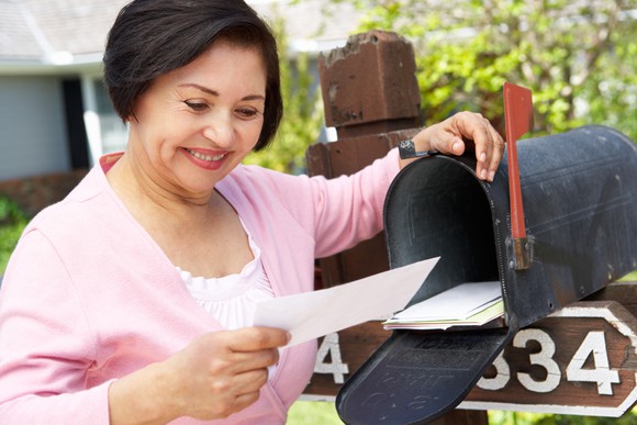 A smiling woman stands next to an open mailbox holding an envelope.